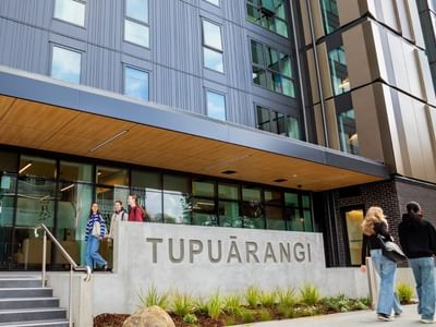 Modern building entrance with concrete Tupuārangi sign, glass doors, and students walking on steps and pathway