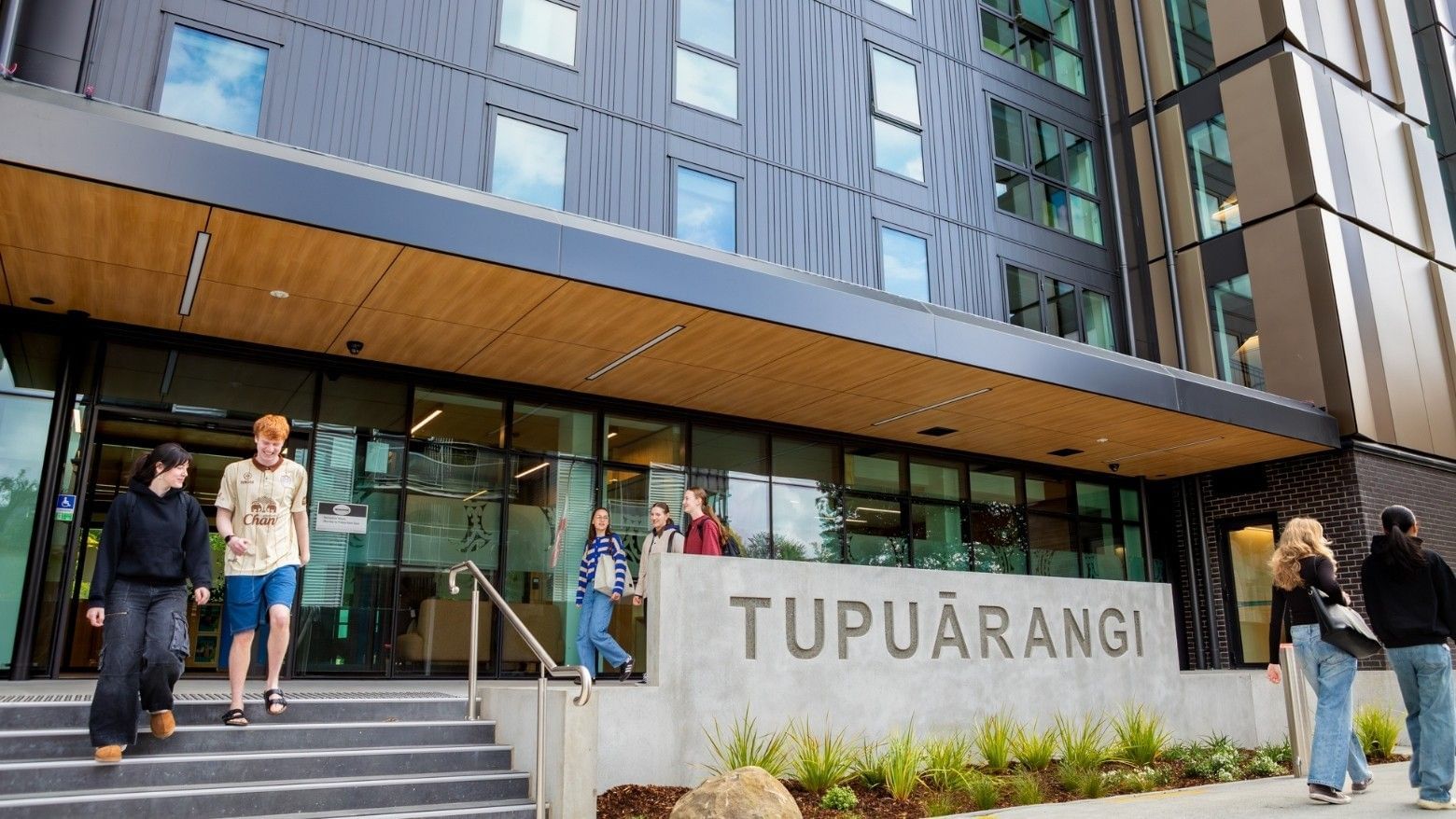 Modern building entrance with concrete Tupuārangi sign, glass doors, and students walking on steps and pathway