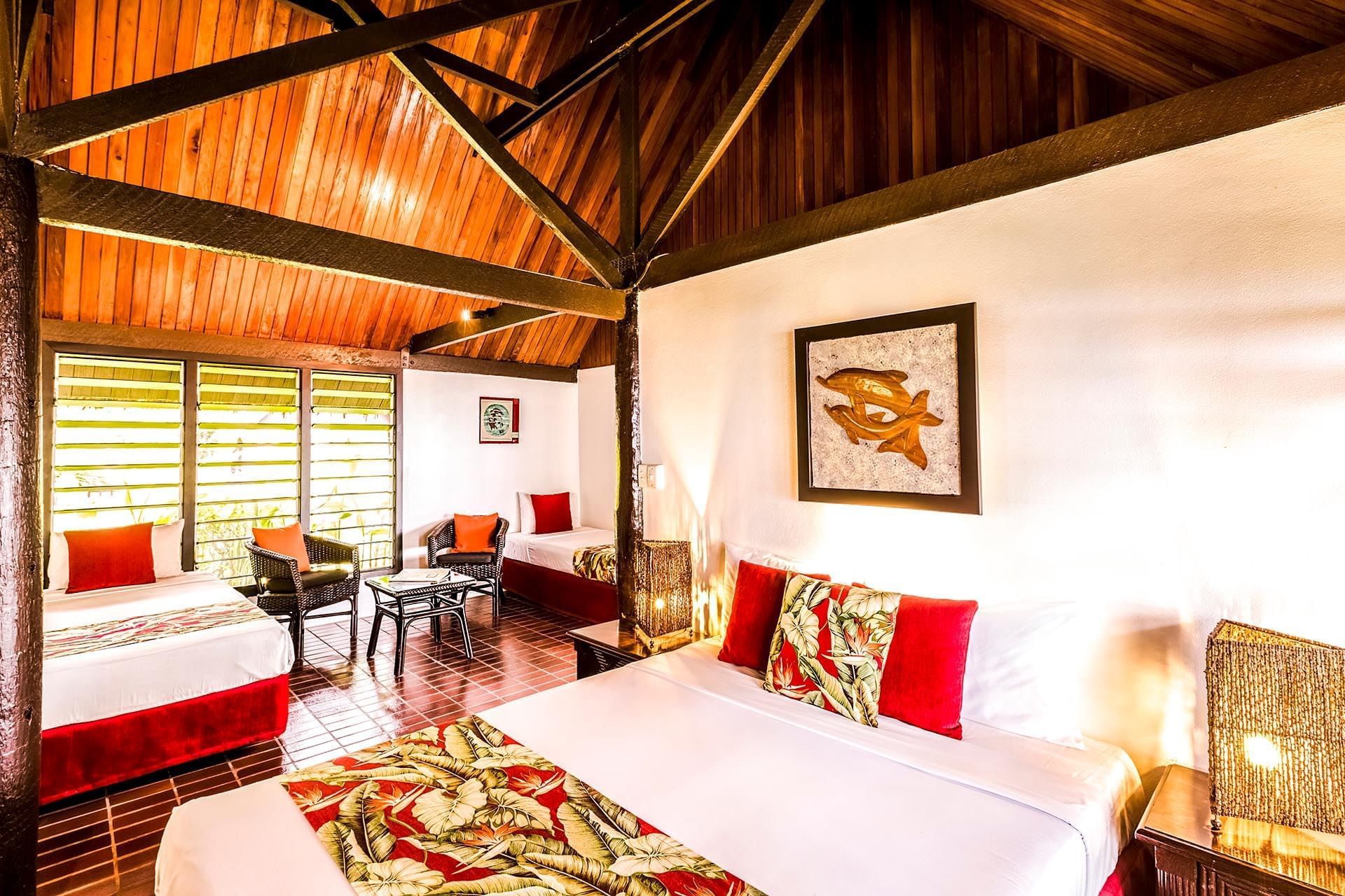 Hotel suite with white beds and red pillows by wicker chairs under a high wood ceiling at Tambua Sands Beach Resort
