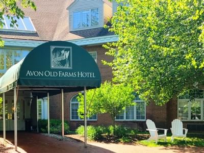 Exterior entrance with green awning and white chairs beside brick building and green trees around Avon Old Farms Hotel