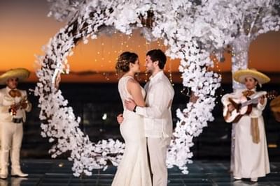 Couple in white attire standing under a white floral arch at sunset with mariachi musicians at Playa Los Arcos Puerto Vallarta 2023