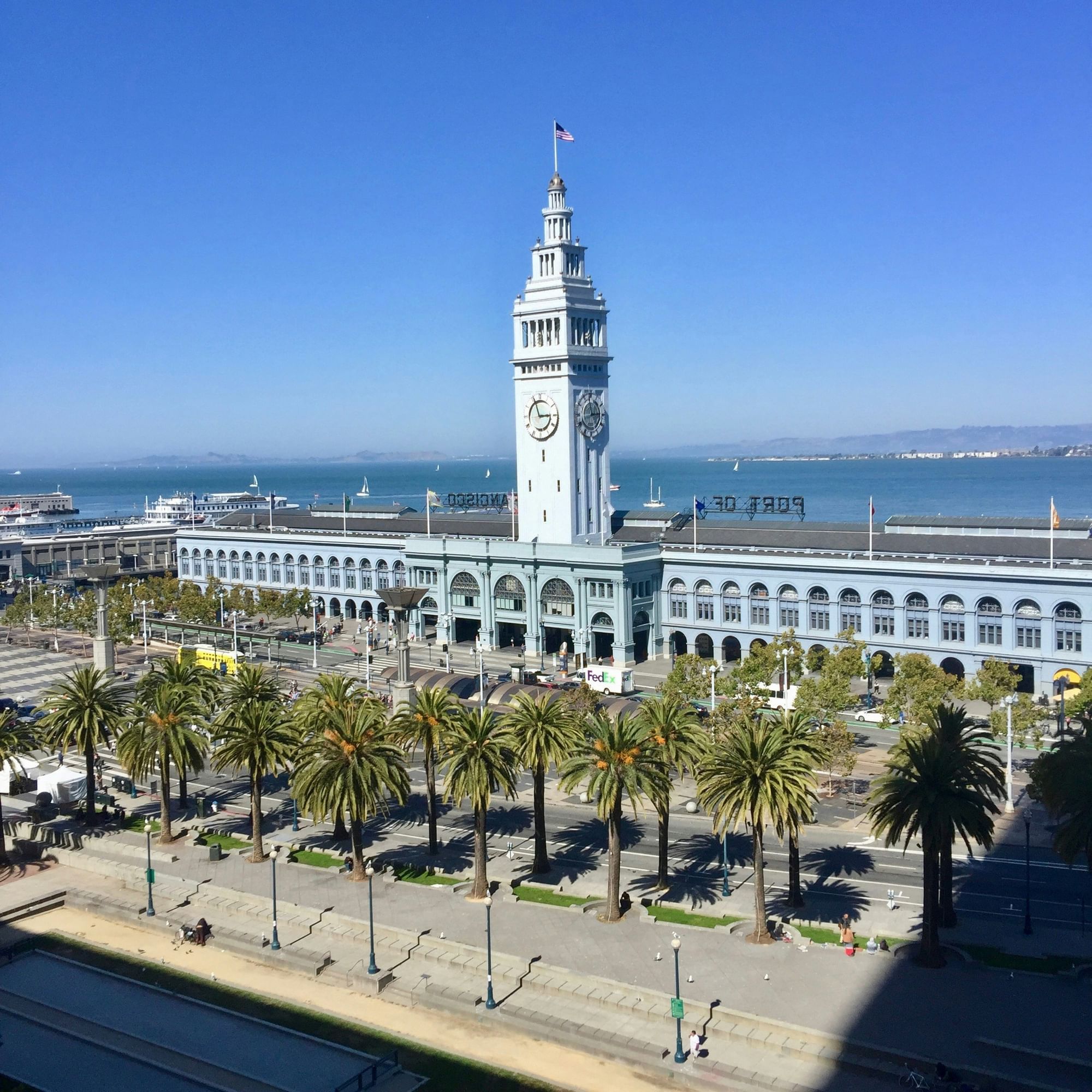 A wide exterior view of a large building with a clock tower, palm trees, and a street with vehicles.