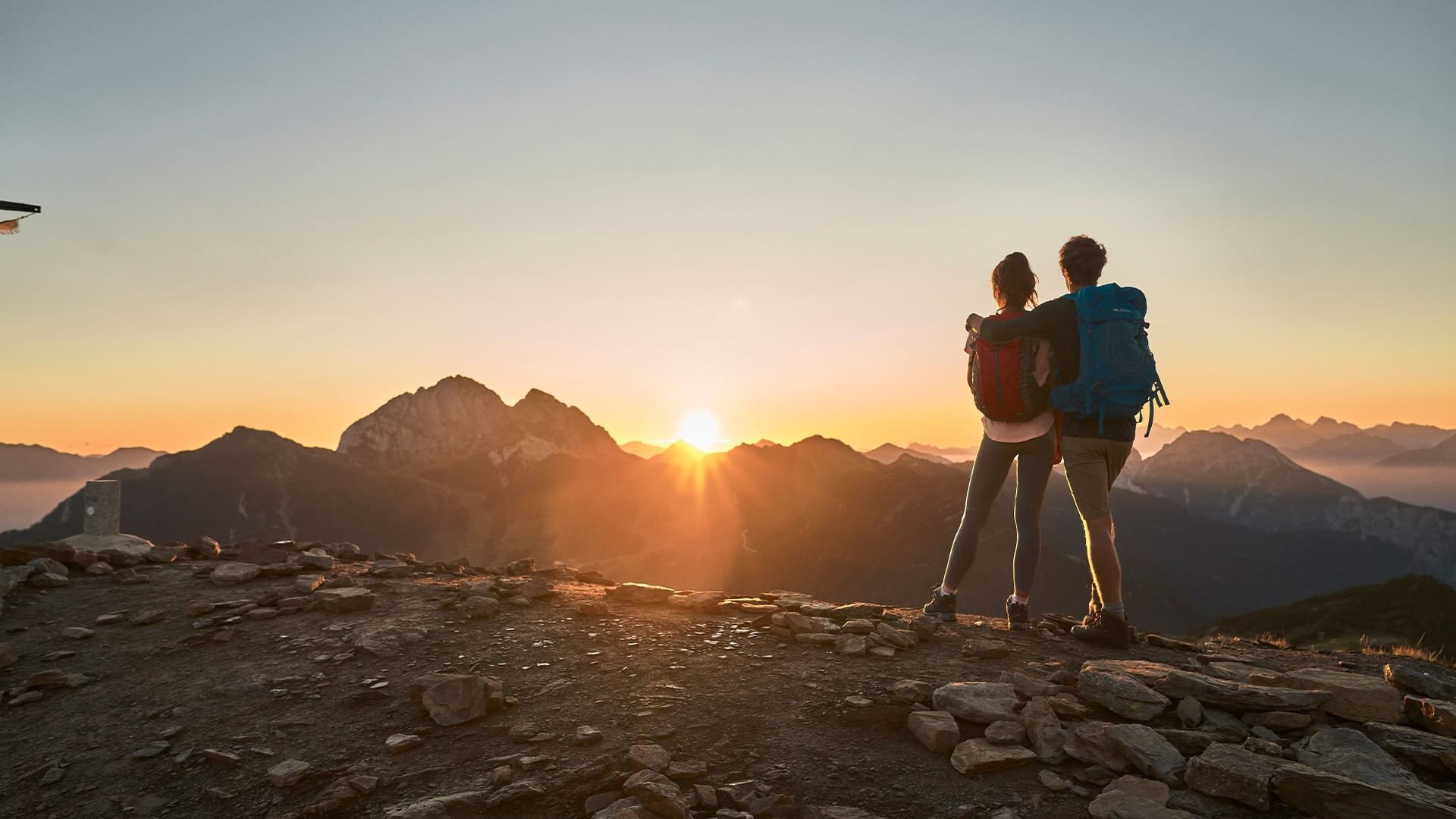 Two hikers with backpacks stand on a rocky mountain peak, watching the sunset over distant mountains.