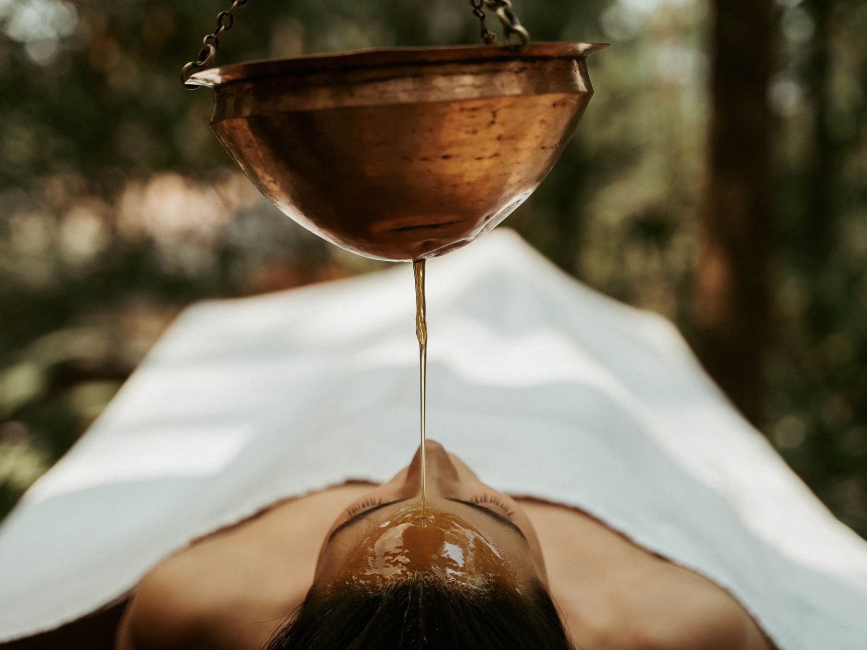Woman on treatment table with warm oil dripping on forehead in a quiet forest setting at Dwarika's Himalayan Wellbeing