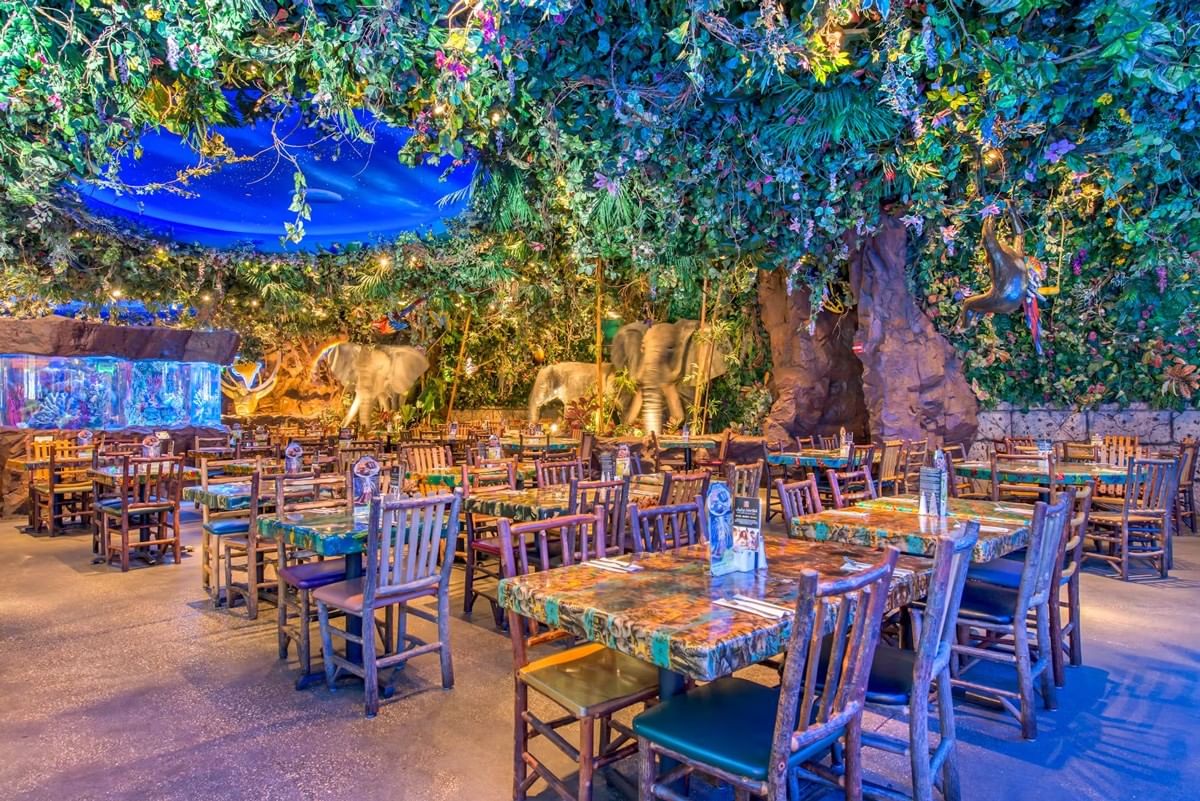 A dining area with wooden chairs and colorful tablecloths, surrounded by greenery and statues.