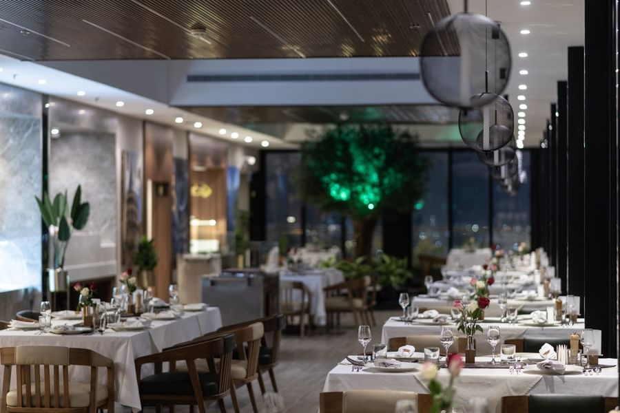 Karkan Restaurant dining area with white tables by glass walls under glowing globe lights at night at Warwick Al Taif Hotel
