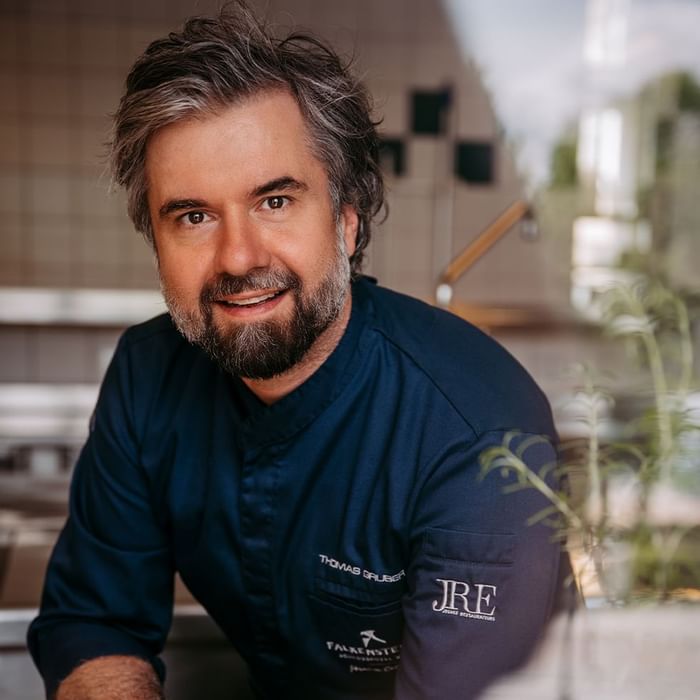 Uomo con barba e capelli grigi sorridente in una cucina, indossa una camicia blu con logo.