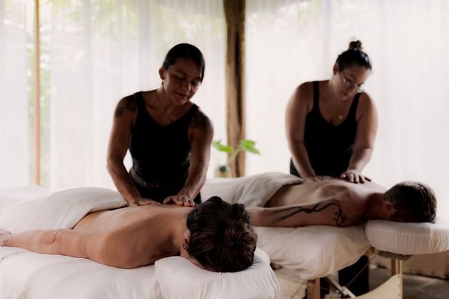 Two people receive back massages in a serene spa setting with white linens and curtains at Cala Luna Boutique Hotel