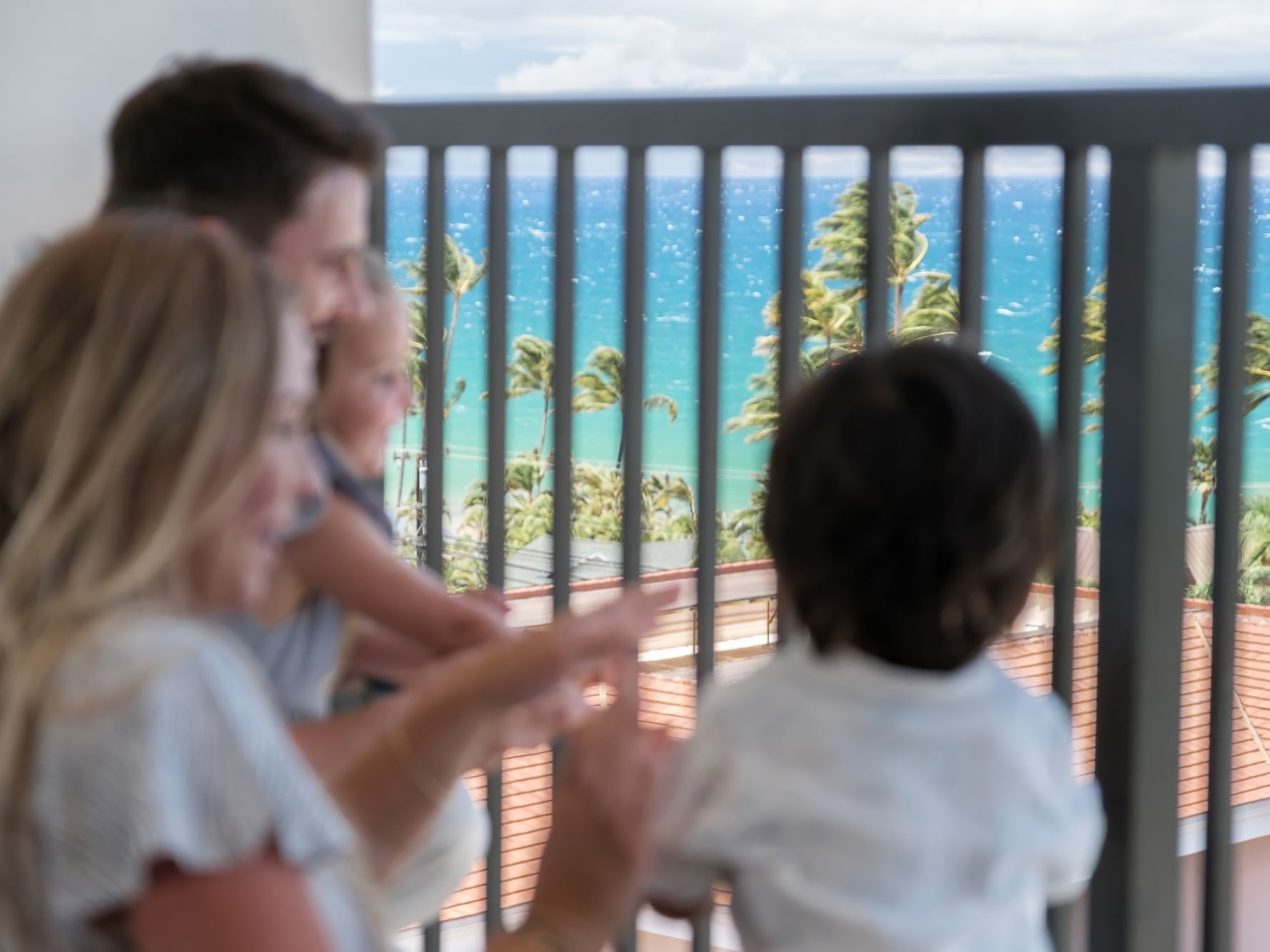 Family with two kids standing on a balcony, admiring the clear blue ocean and palm trees at the Maui Coast Hotel