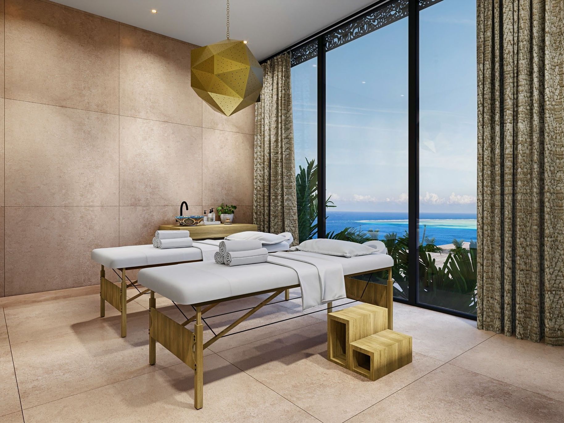 Two massage tables with towels and pillows in a room with ocean view through large windows.