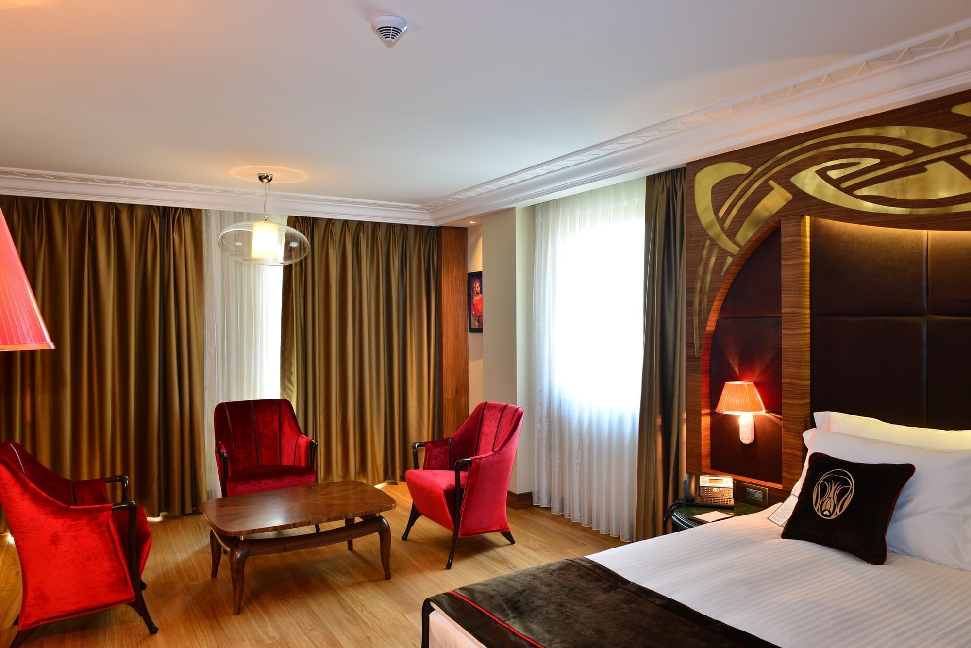 Red chairs by a coffee table surrounding a plush bed under a gold headboard in Junior Suite at Warwick Ankara