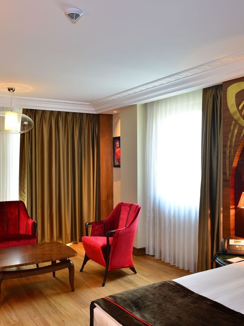 Junior suite with Plush bed by red velvet chairs surrounding a coffee table under a bright window at Warwick Ankara