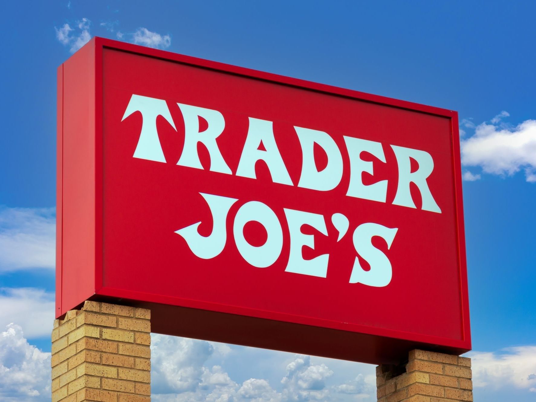 Red Trader Joe's sign mounted on a brick pillar against a blue sky with clouds near Tradewinds Apartment Hotel