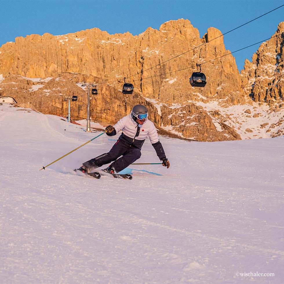 Sciatore in discesa su pista innevata con montagne rocciose e seggiovie sullo sfondo.