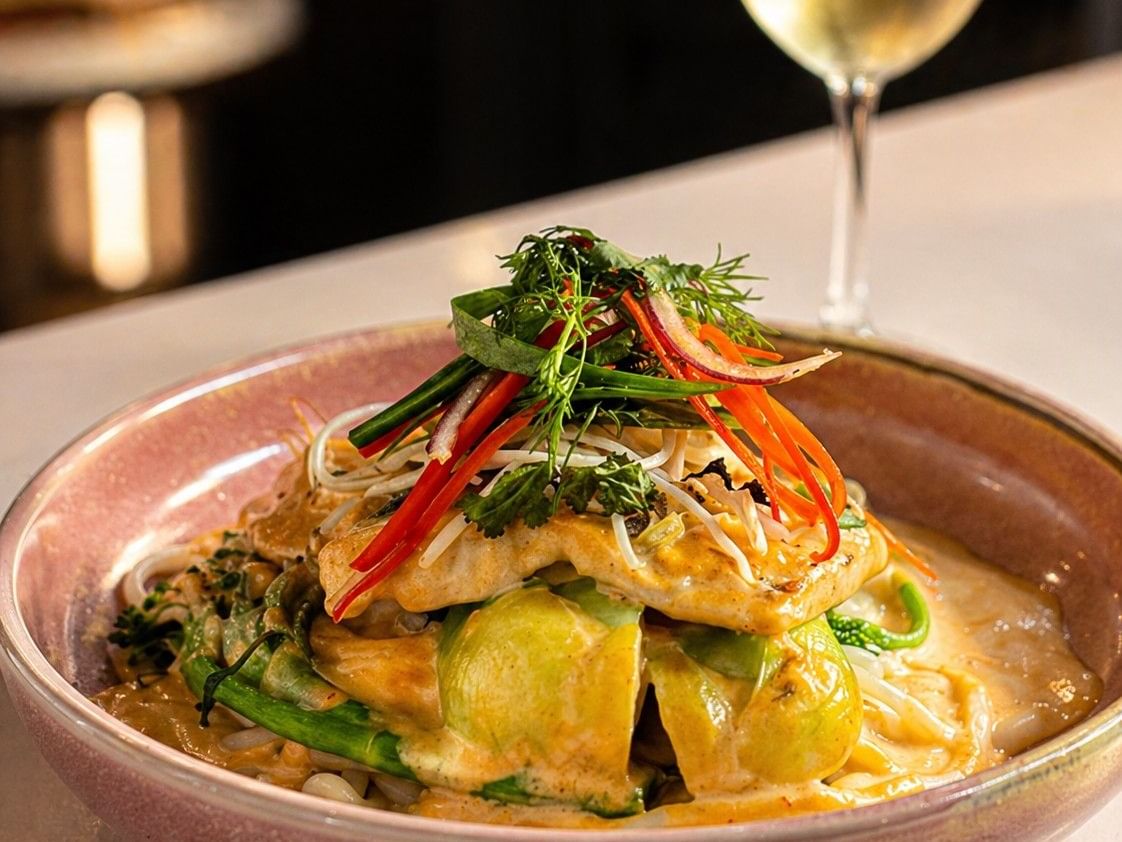 A fragrant fish curry served over thick noodles and asian greens, topped with a fresh herb salad next to a glass of white wine.