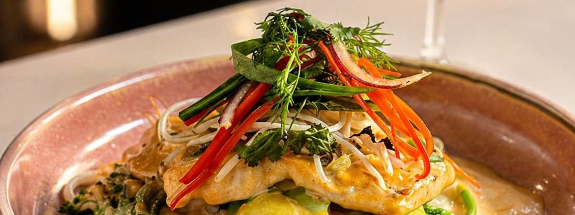 Pan-seared fish served over noodles and seasonal vegetables in a creamy sauce, garnished with fresh herbs and accompanied by a glass of white wine.