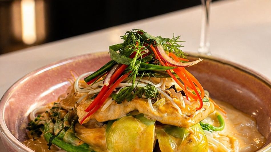 Pan-seared fish served over noodles and seasonal vegetables in a creamy sauce, garnished with fresh herbs and accompanied by a glass of white wine.