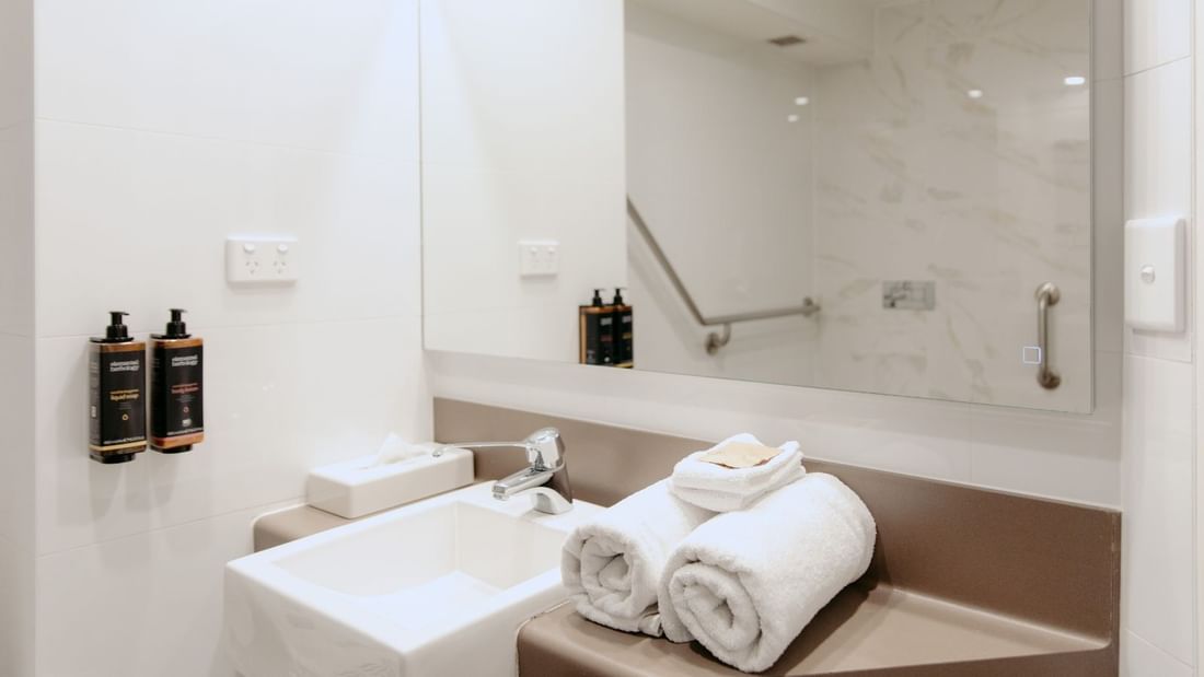Bathroom with white sink, rolled towels, and a mirror reflecting a handrail and outlet.
