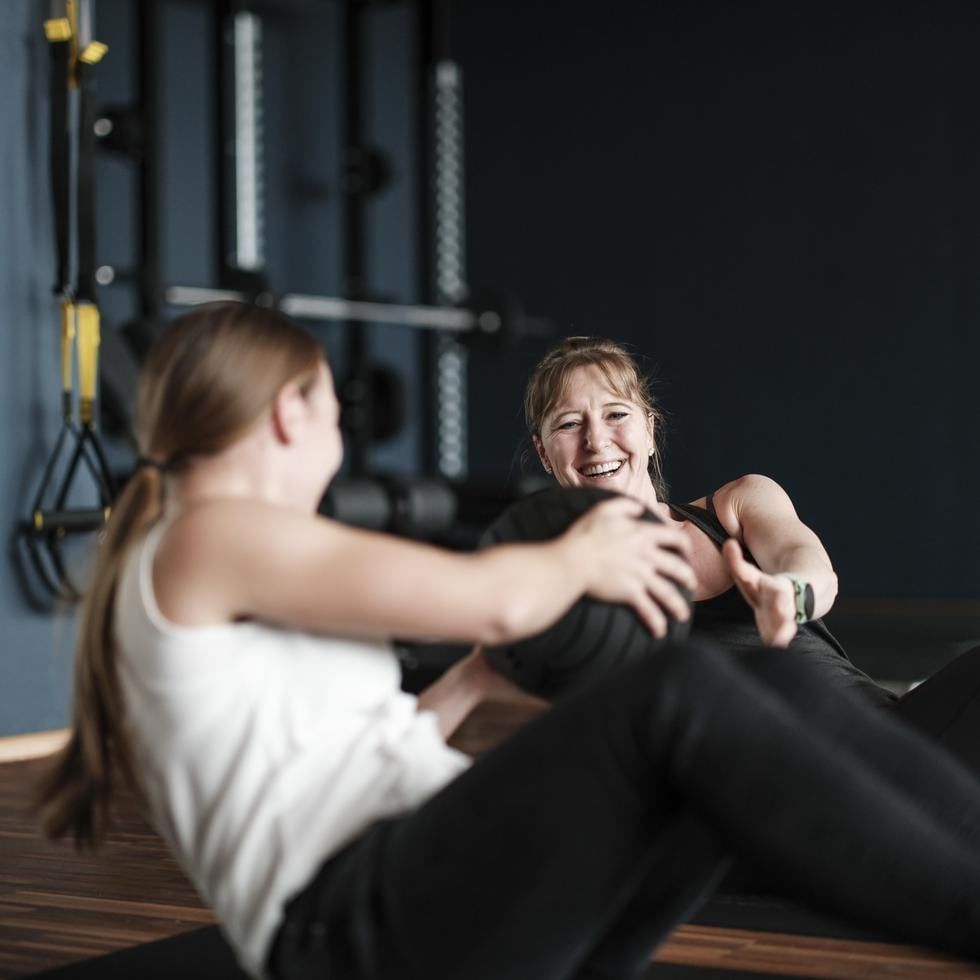 Two women in a gym, one instructing the other on using a medicine ball.