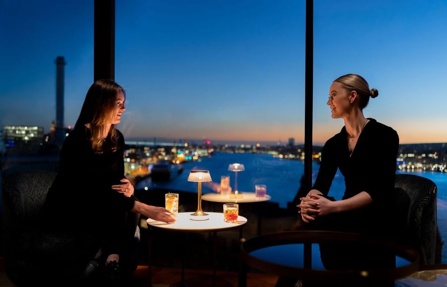 Two women in black shirts sit at a table with drinks and lamps, overlooking a city at dusk.