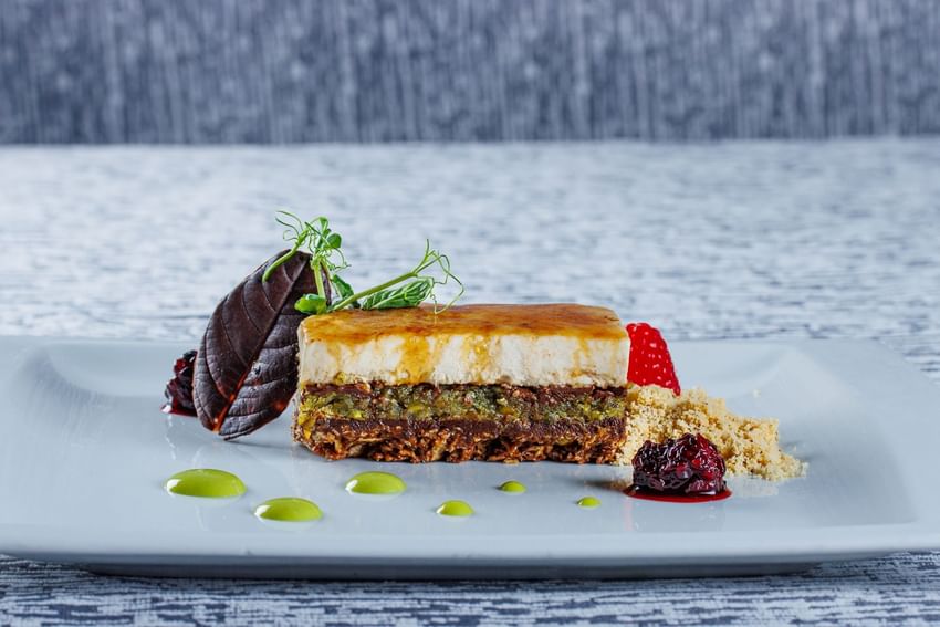 Primer plano de un postre gourmet de tres capas con un acabado brillante, nueces y crema servido sobre una mesa en el Sunset Plaza Beach Resort