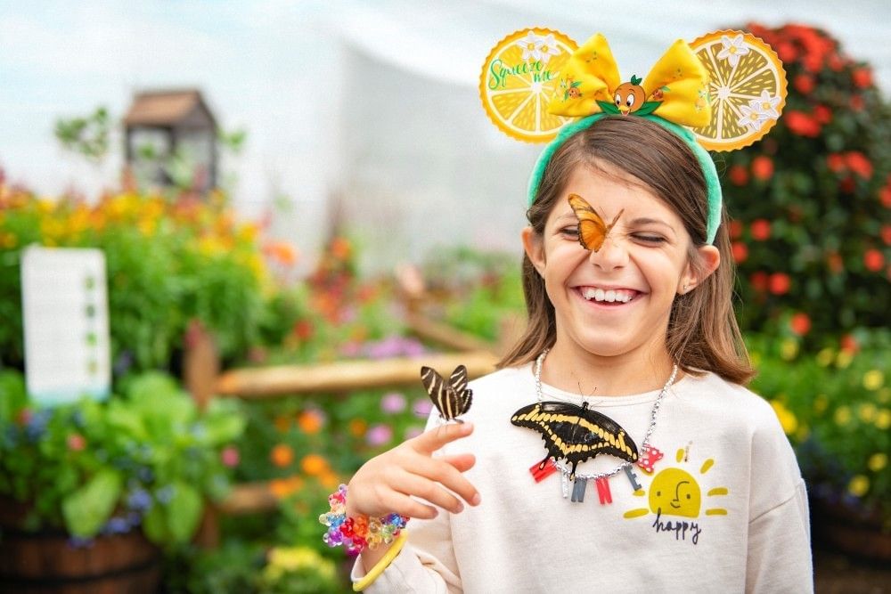 A girl in citrus themed Minnie Mouse ears smiles in a garden with butterflies on her nose, finger, and chest.