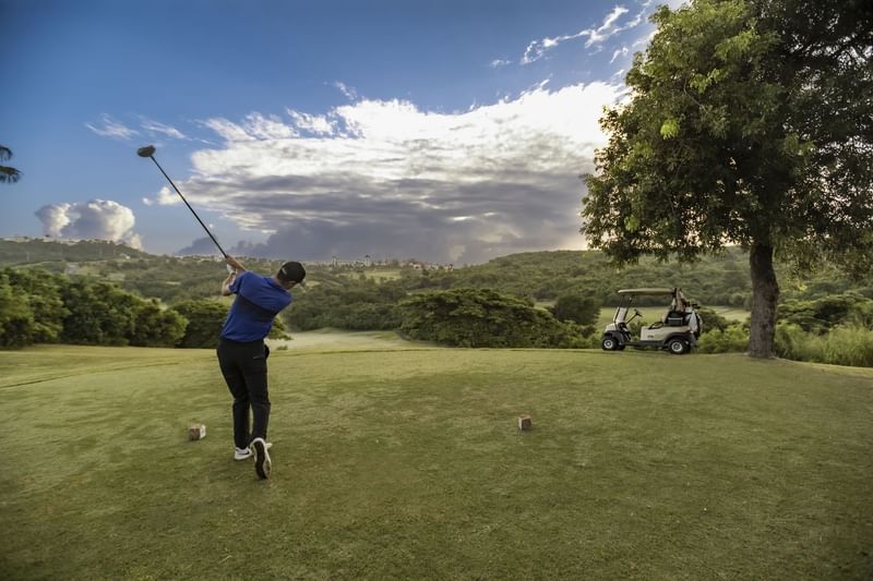 Golfer taking a swing on a scenic green course next to a golf cart parked under a large tree at El Conquistador Resort