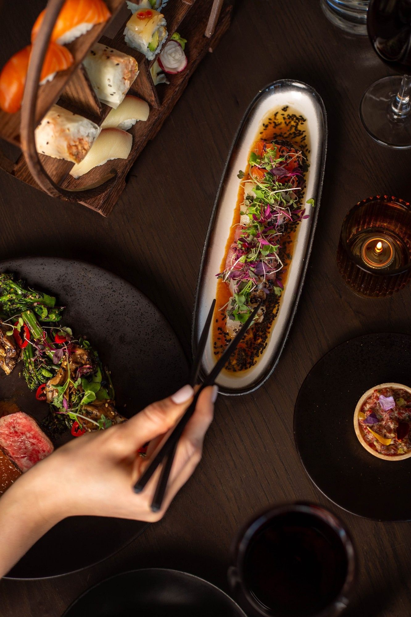 Hand holding chopsticks reaching Sushi and a plate of seared meat with garnishes in Valerie at Hotel X Toronto