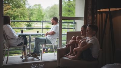 Family of four in a hotel room with two kids on a sofa and parents on a balcony.