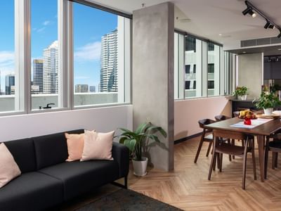 Urban lounge interior with large windows, black sofa with pink cushions, wooden dining table, and cityscape view.