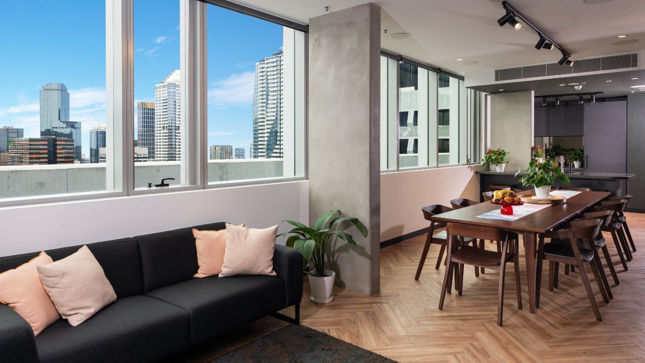 Urban lounge interior with large windows, black sofa with pink cushions, wooden dining table, and cityscape view.
