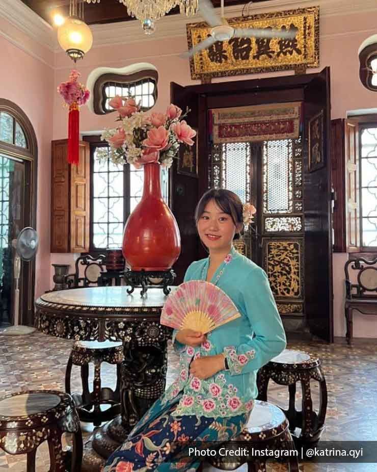 A lady dressed in Nyonya Kebaya sat on a chair while holding a fan. - Lexis Suites Penang