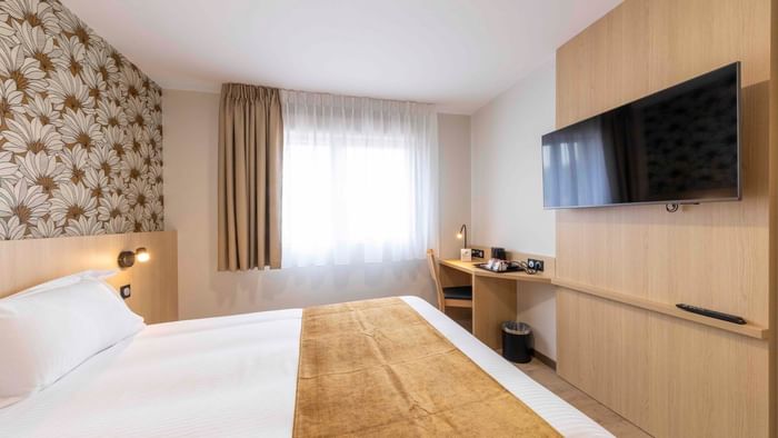 A cozy hotel room with a bed, wooden desk, flat-screen TV, and window at The Originals City Hotel du Parc Gravelines.