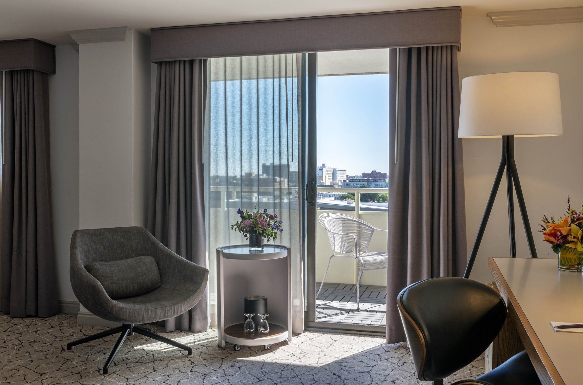 A hotel room with a desk, chair, lamp, and balcony with a city view.
