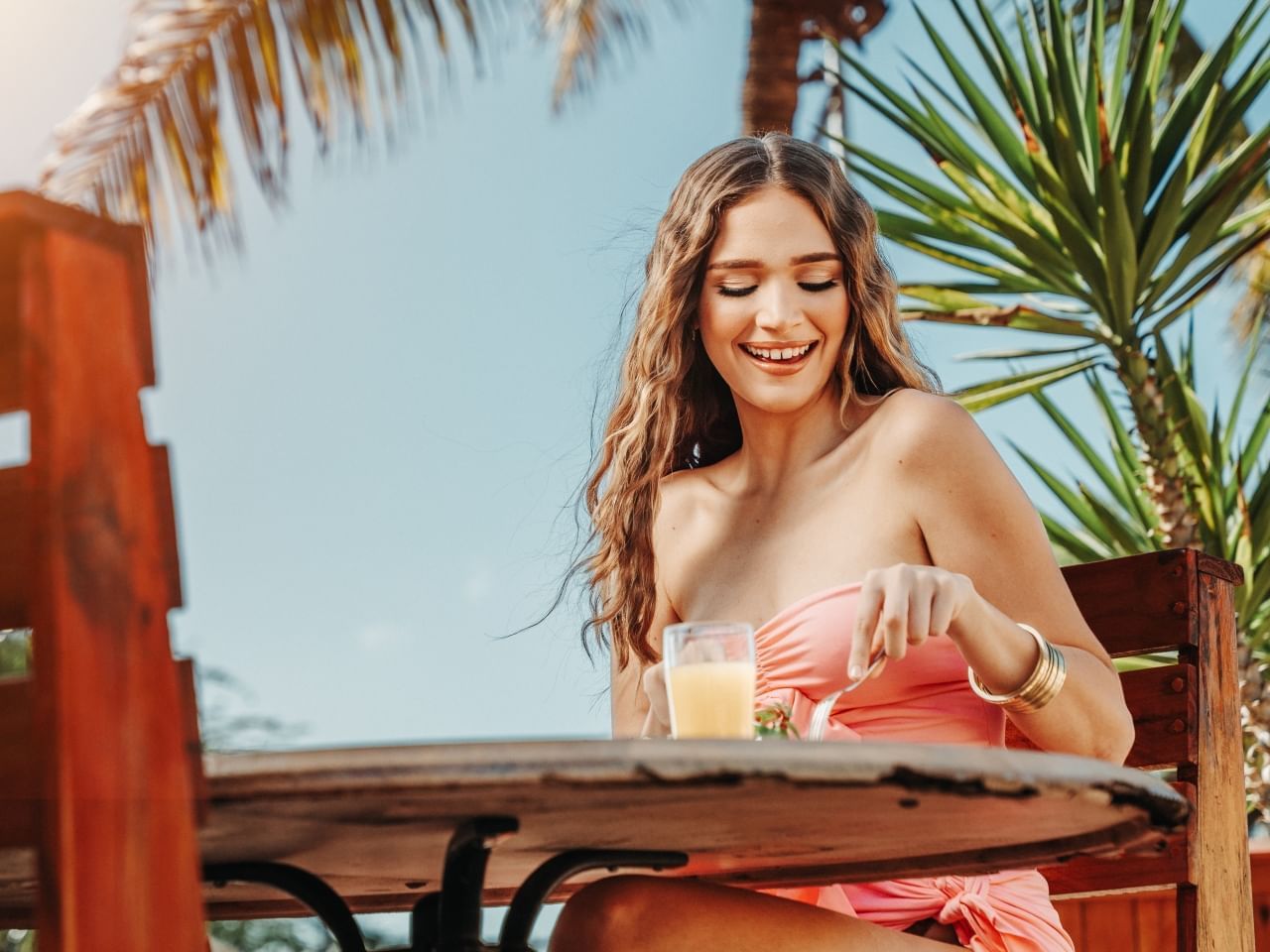 Woman in pink dress smiling while sitting and eating at a table in a tropical setting.