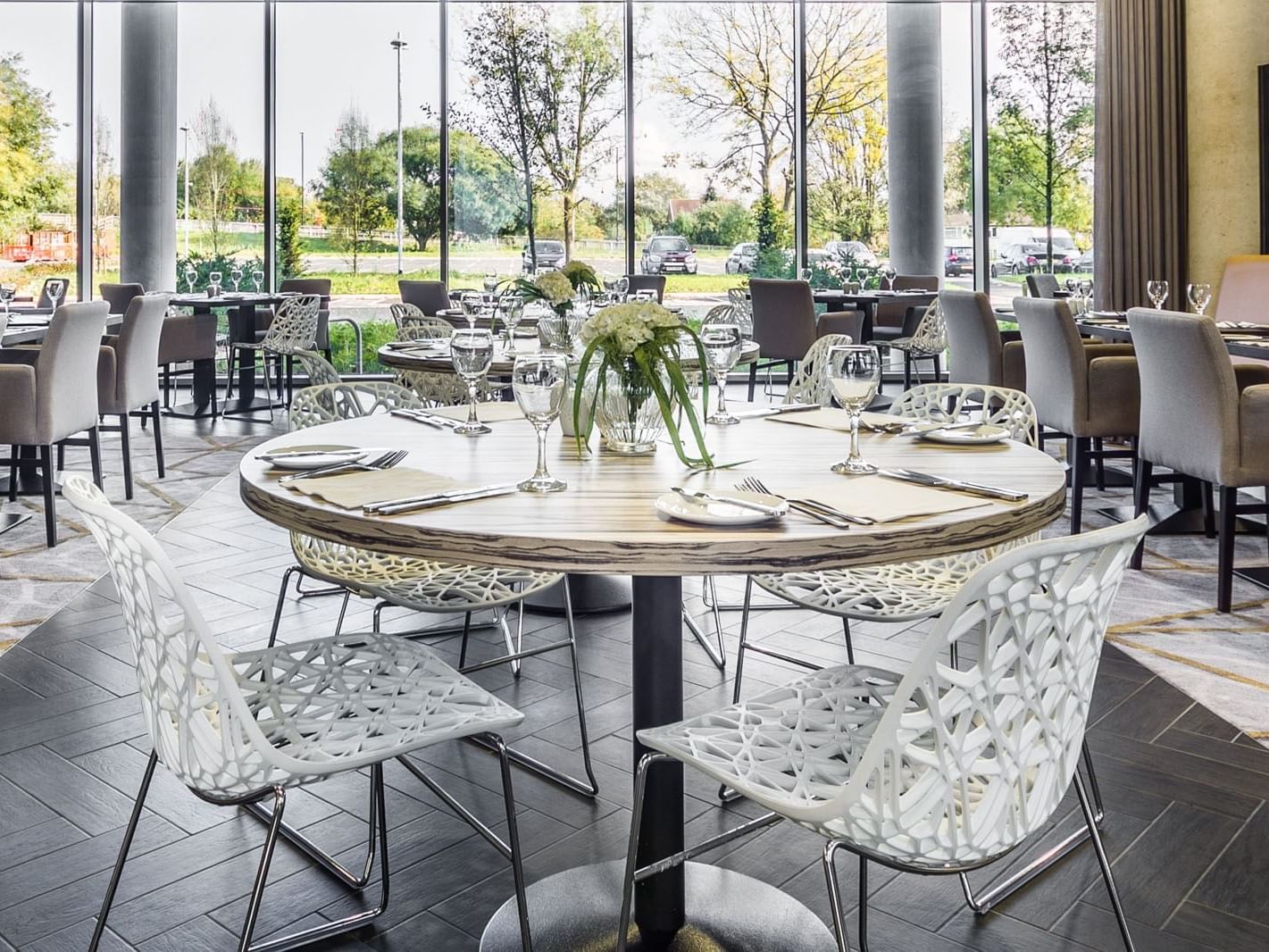 Glass House Restaurant with round tables and stylish chairs by large windows facing greenery at Heston Hyde Hotel