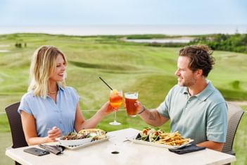 Couple raising glasses in a toast over a patio lunch with green hills behind Rodd Crowbush Golf & Beach Resort