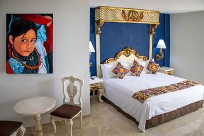 Hotel room with white bed, blue ornate headboard, colorful artwork, and decorative pillows at Playa Los Arcos Puerto Vallarta 2023