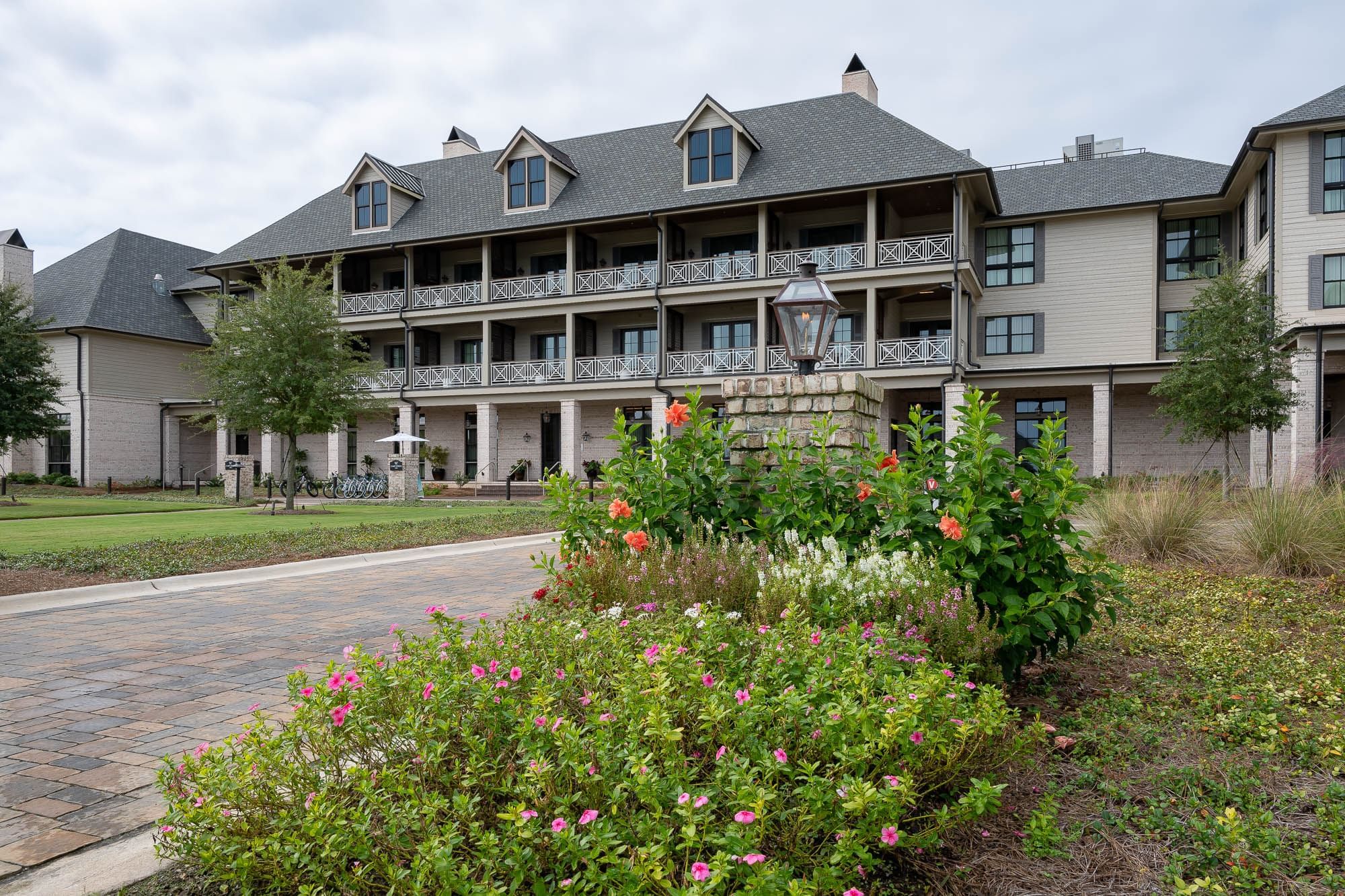 Large building with gray roof and balconies, surrounded by flowers and greenery under a cloudy sky.