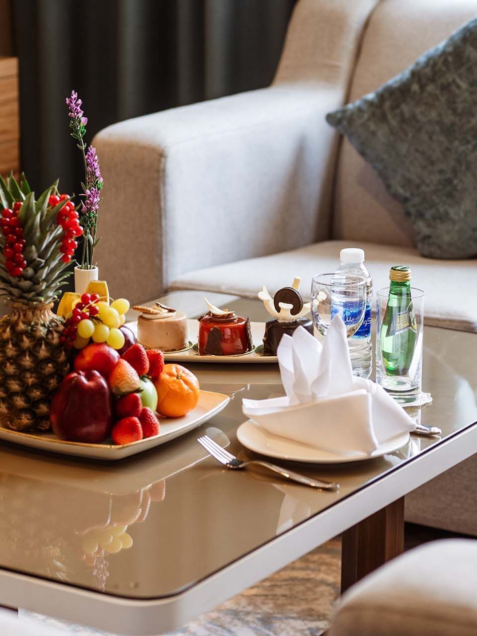 Fruit platter and pastries on a glass table by a grey sofa in Ambassador Suite at Cantonal Hotel by Warwick Riyadh