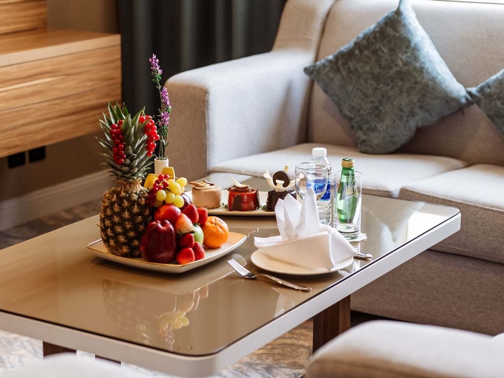 Fruit platter and pastries on a glass table by a grey sofa in Ambassador Suite at Cantonal Hotel by Warwick Riyadh