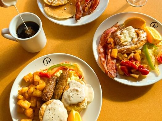 Three breakfast plates with eggs, bacon, pancakes, and hash browns alongside a cup of coffee on a golden table.