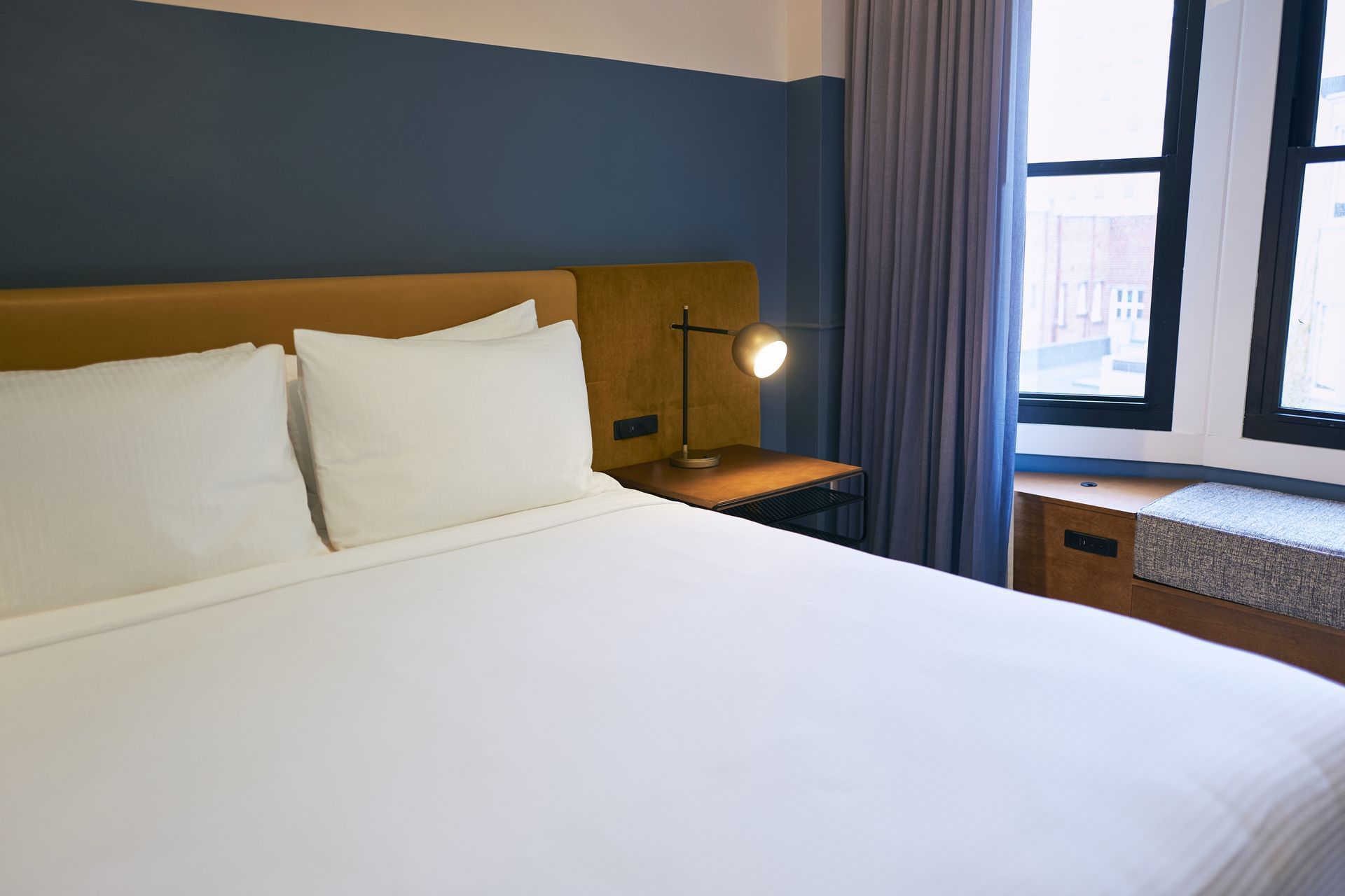 A neatly made bed with white linens and pillows against a blue and brown headboard, with a reading lamp and window seat beside it.
