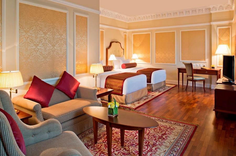 Junior Suite features Gray sofas with red pillows by an oval coffee table surrounding twin beds at Warwick Doha Hotel