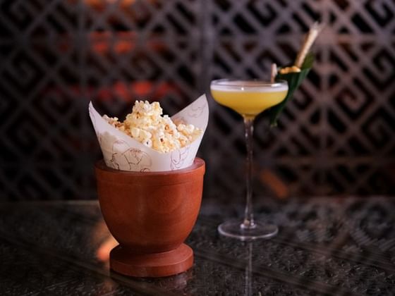 Clay pot of popcorn next to a yellow cocktail with a leaf garnish served in 2 Lam Son at Park Hyatt Saigon