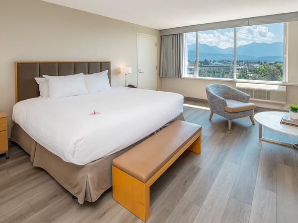 A hotel room with a large king bed, nightstand, chair, round table, and large windows with mountain view.