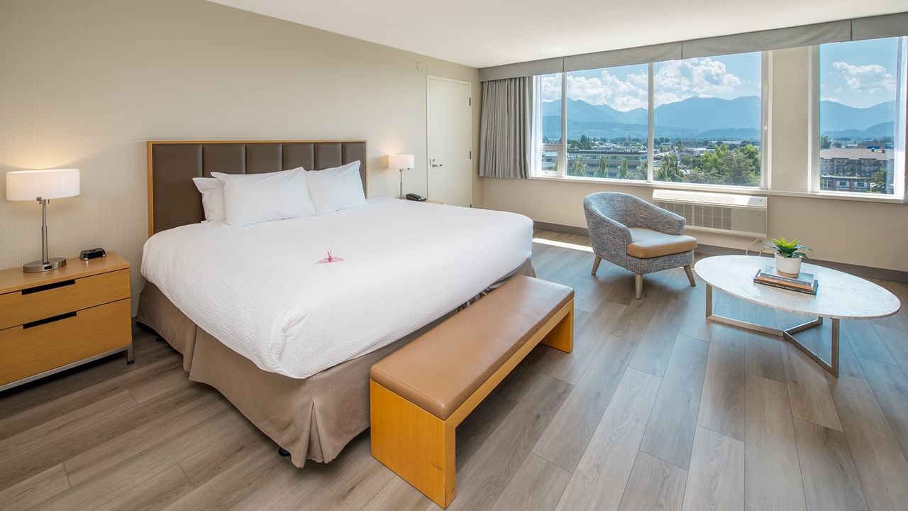 A hotel room with a large king bed, nightstand, chair, round table, and large windows with mountain view.