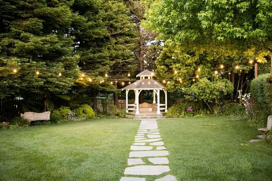 Outdoor gazebo with string lights above a stone path in a garden with green lawn and surrounding trees