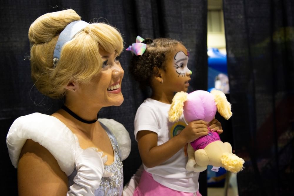 A woman dressed as Cinderella smiles at a camera beside a little girl with face paint holding a stuffed toy at the Florida Kids and Family Expo..