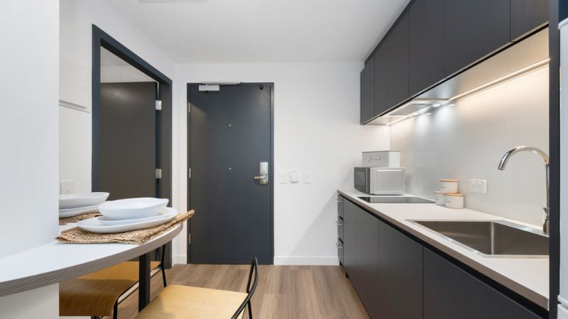 Modern kitchen with black cabinets, white counters, sink, microwave, and dining area with wooden table and chairs.